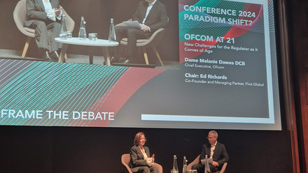 Photo of a woman and man sat on stage, on chairs with a small table between them and a large screen behind them with their image projected onto it.