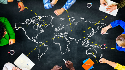 Overhead photo of a blackboard desk with a map of the world drawn in white and markers in yellow. People sit around the edge.