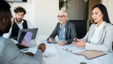 A small group of diverse individuals sat around a desk with paperwork showing charts and graphs. They look engaged.