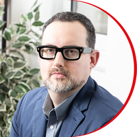 Headshot: Ross Lewis, a man with short dark hair and beard, wearing dark, thick-rimmed glasses and a dark blue jacket.