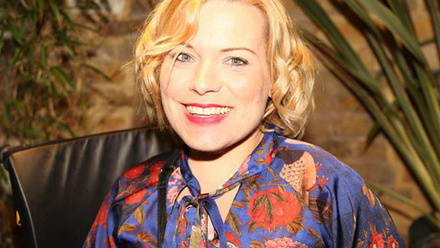 Headshot: A woman with short curly blonde hair wearing a colourful patterned dress.