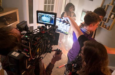 Production image showing a camera person holding a large broadcast camera with a clapper and three actors in front of it.