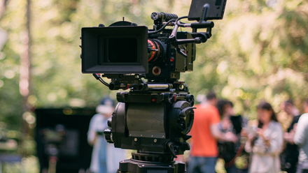 Photo of a broadcast camera on a tripod in a forest. A crew of people can be seen in the background.