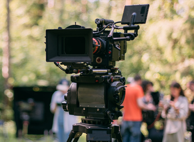 Photo of a broadcast camera on a tripod in a forest. A crew of people can be seen in the background.