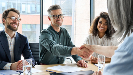 Three people sit opposite a woman with long grey hair, they are smiling and the person in the middle is shaking hands with her over a desk.