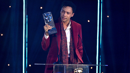 Actor, Kit Young dressed in a claret suit and white shirt is holding the Actor Film Award up whilst stood behind a podium with the BAFTA logo on it.