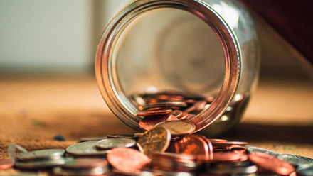 Photo close up of a tipped over glass jar with coins spilling out of it.