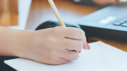 Photo close up of a hand holding a pen over a piece of paper. A laptop mouse and keyboard can be seen in the background.