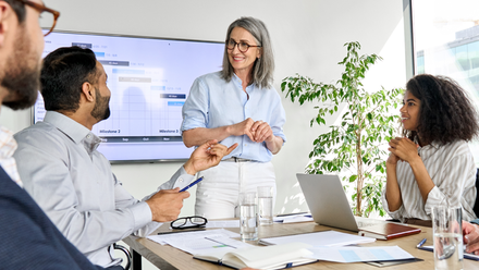 A diverse group of professionals in a meeting room, with a mature woman standing and leading a discussion.