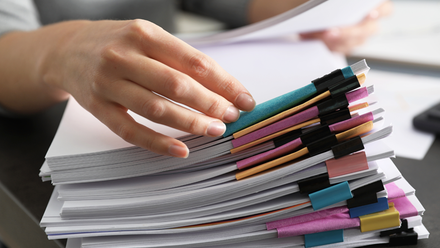 Photo close up on a stack of documents with multi-coloured post it notes clipped to the tops of them, and somebody's hands sorting through them.