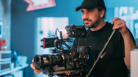 Photo of a man stood behind a camera with his hand on the tripod grip and lens. He is wearing a cap and black t-shirt.