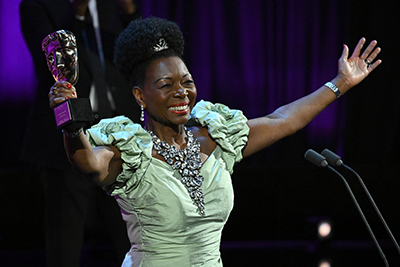 Photo: Dame Floella Benjamin wearing a light green formal dress, holds her BAFTA Award aloft and smiles brightly towards the audience out of frame.