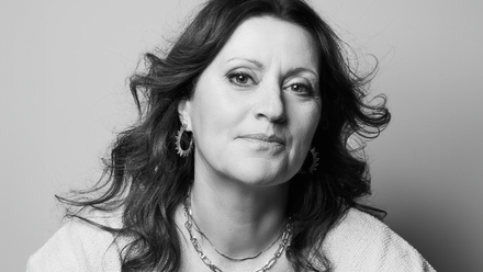 Headshot: A woman with long wavy brown hair wearing a white top and necklace.