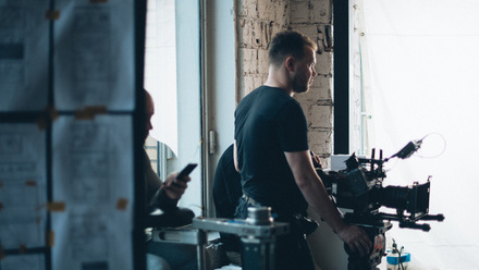 Photo of a man on set, stood behind a camera. Another man is sat behind him and is on his phone. Papers attached to a board seen in the foreground.