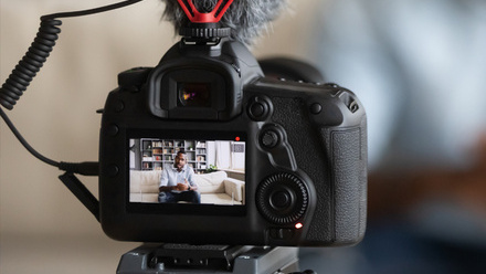 Photo of a DLSR video camera from behind, in the viewfinder we can see a subject sitting on a sofa with bookshelves behind him. 