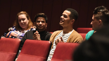 Close up photo of four diverse individuals sat in a row in a cinema space, one of whom has raised hands and is making a point.