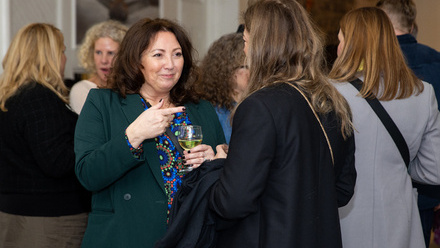 A woman holding a glass of wine engages in conversation with another attendee at a lively networking event.