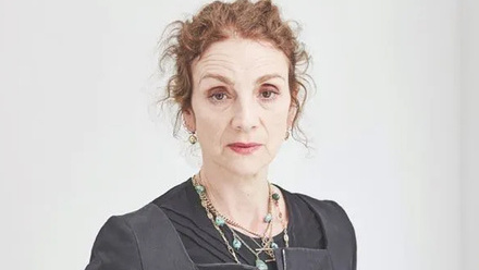 Headshot: Sally Woodward Gentle, a woman with wavy auburn hair tied back wearing a black top and jacket and a gemstone necklace.