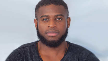 Headshot: Josh Wilson, a man with short black afro hair and a beard, wearing a dark-striped t-shirt.