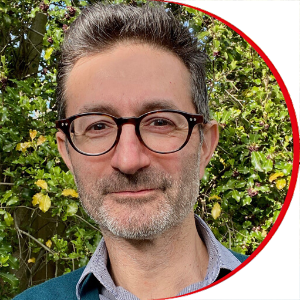 Headshot: Paul Cohen - a man with short black hair and a greying short beard wearing black-rimmed glasses and a light blue shirt.