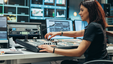 Photo of a woman at a production control desk viewing a bank of screens and adjusting settings.