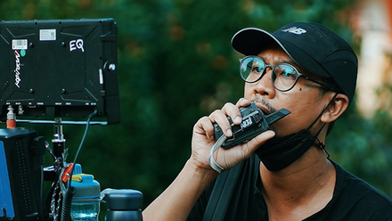 Photo of a director on location, he is standing behind a monitor and holding a walkie-talkie up to his mouth.