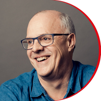 Headshot: Billy Macqueen, a man wearing black-rimmed glasses and a denim shirt, looking and smiling off to left of frame.