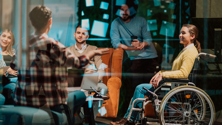 Photo of a group of office workers in a breakout room, including a woman in a wheelchair who is smiling at the person speaking.