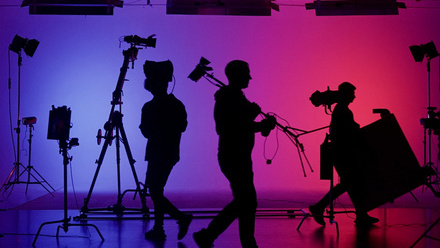 Silhouette of various crew members on a set, moving equipment around. The background it lit in vibrant purple and pink.