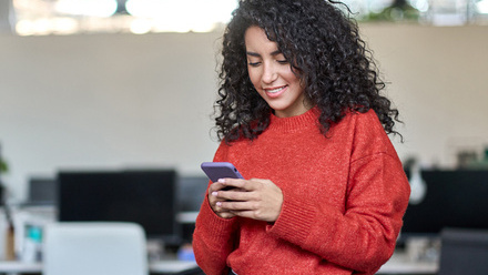 Photo of a woman holding a smartphone in her hands and looking down at it, stood in an office environment.