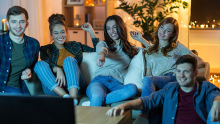 Five young adults are sat around the TV, watching intently and looking engaged and entertained, basked in blue light.