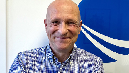 Headshot: Alan Clement, a man with a bald head wearing a blue and white checked shirt.