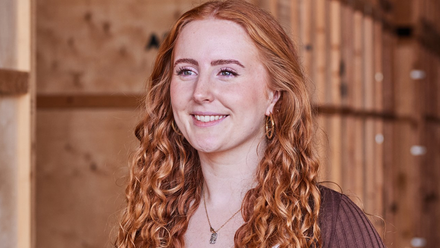 Headshot: A young woman with long curly red hair wearing a white top and necklace.