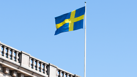 Photograph of the Swedish flag being flown on a mast above a traditional-looking brick building, against a clear blue sky.