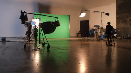 Photo of a large studio with a green screen and lights set up within it and a couple of crew members.